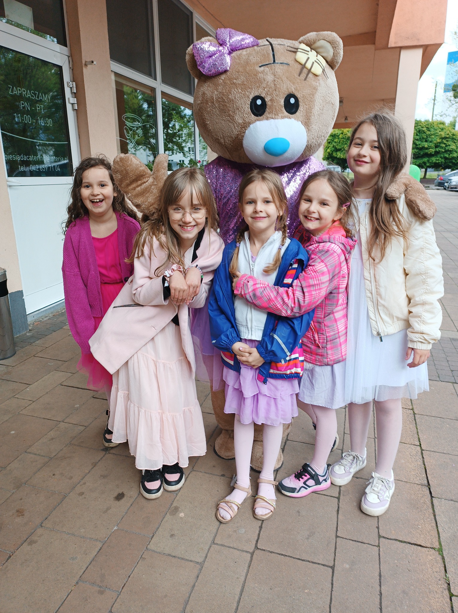 Uśmiechnięte dziewczynki pozują z animatorem przebranym za dużego misia z fioletową kokardą. Dziewczynki ubrane w różowe, białe i fioletowe sukienki i kurtki.