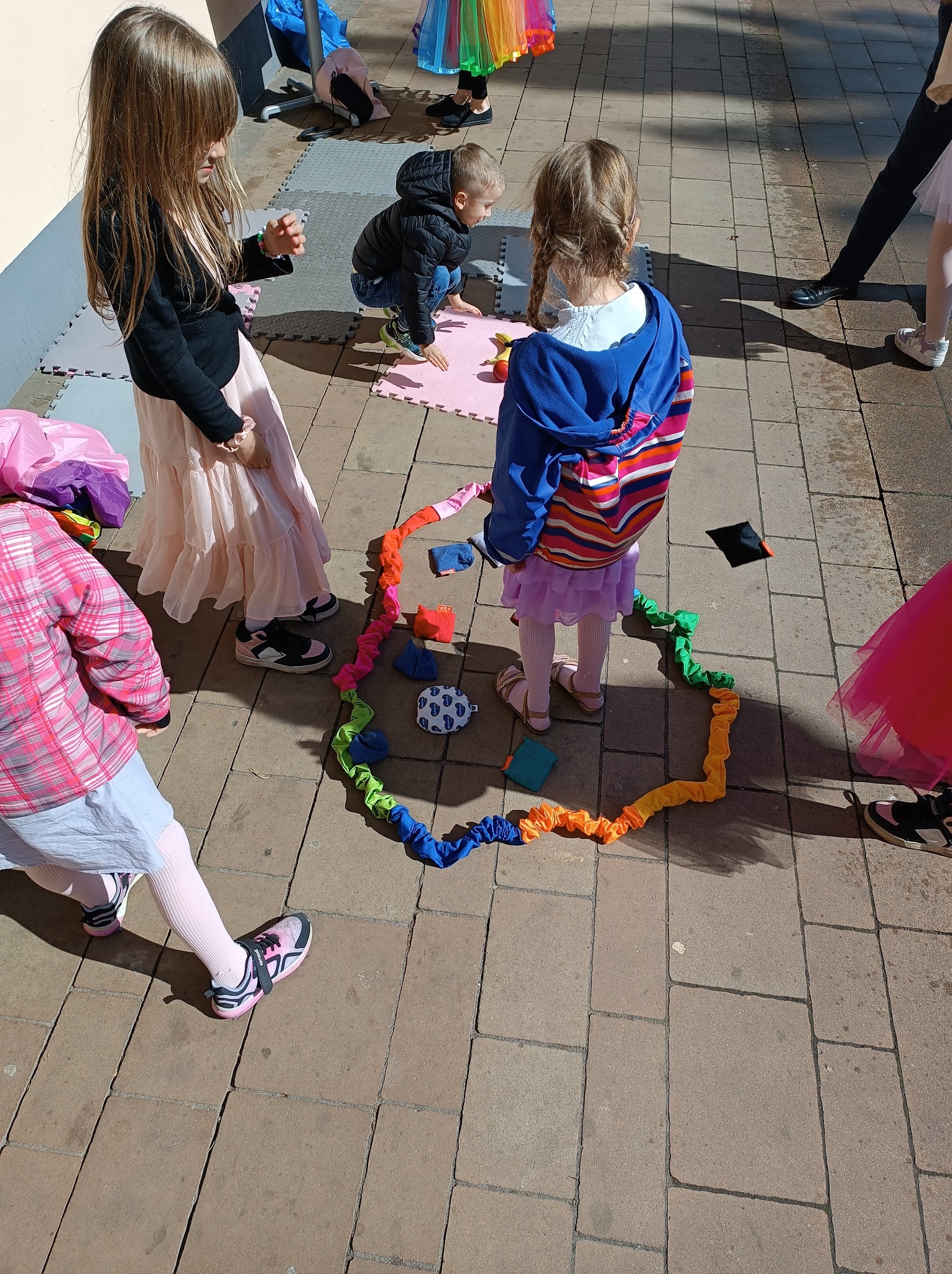 Dzieci bawią się na zewnątrz, układając kolorową, elastyczną linę w kształt serca i rozsypując wokół niej małe, miękkie woreczki z wypełnieniem.