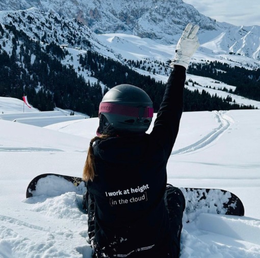 Osoba w kurtce z napisem 'I work at height in the cloud' siedzi na śniegu z deską snowboardową, góry w tle. Nietypowy napis na odzieży.