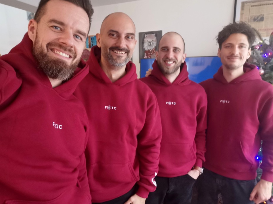 Grupa uśmiechniętych mężczyzn we Wrocławiu prezentuje bordowe bluzy z kapturem, z małym, białym logo na piersi. W tle widoczna choinka.