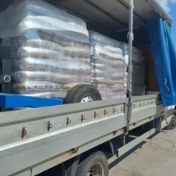 Załadunek pelletu w workach na pakę samochodu dostawczego z plandeką. Widoczny fragment podwozia i koła zapasowego. Słoneczny dzień.