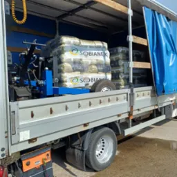 Otwarta paka dostawczego auta z załadowanym pelletem marki Sobianek, zabezpieczonym na niebieskim stelażu. Widoczne narzędzia i opona. Częściowo zasłonięte niebieską plandeką.