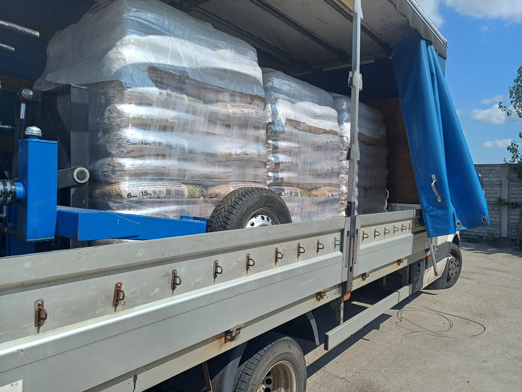 Załadunek pelletu w workach na pakę samochodu dostawczego z plandeką. Widoczny fragment podwozia i koła zapasowego. Słoneczny dzień.