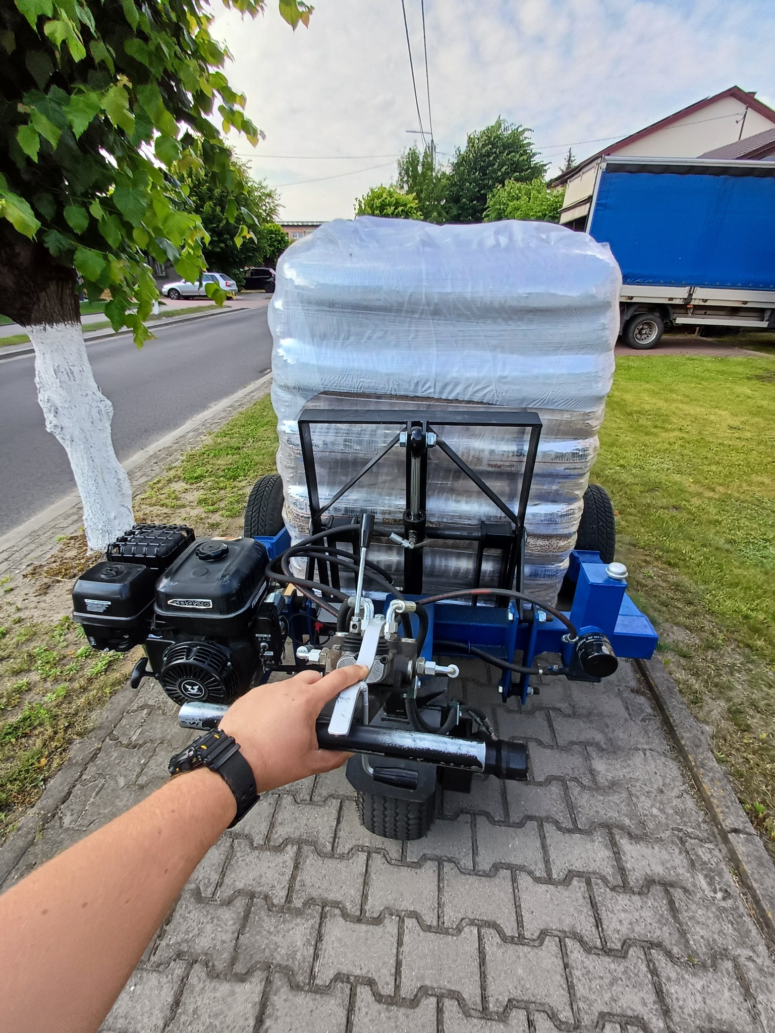 Wózek transportowy z silnikiem spalinowym przewożący paletę z workami, prawdopodobnie z pelletem. Widoczny fragment chodnika, trawnika i niebieska plandeka samochodu ciężarowego.