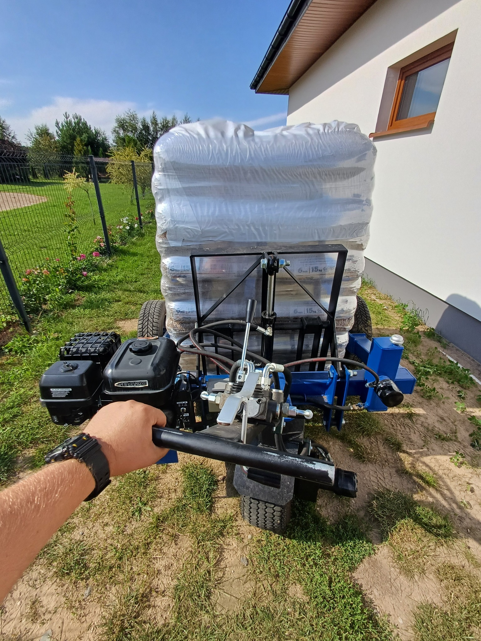 Maszyna do pakowania pelletu z załadowanymi workami 15kg, widok z perspektywy operatora, na tle zielonego trawnika i białego budynku z oknem.