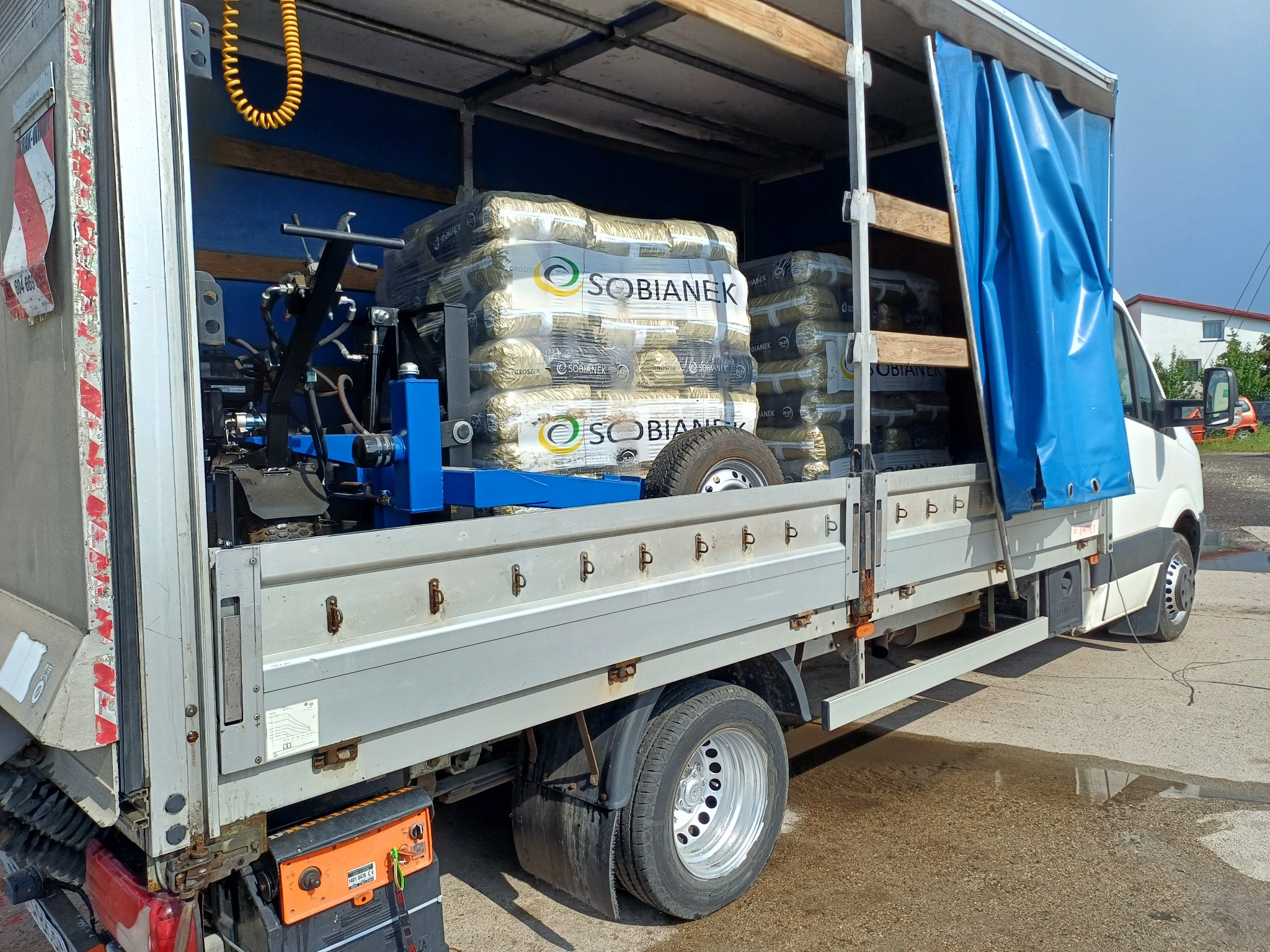 Otwarta paka dostawczego auta z załadowanym pelletem marki Sobianek, zabezpieczonym na niebieskim stelażu. Widoczne narzędzia i opona. Częściowo zasłonięte niebieską plandeką.