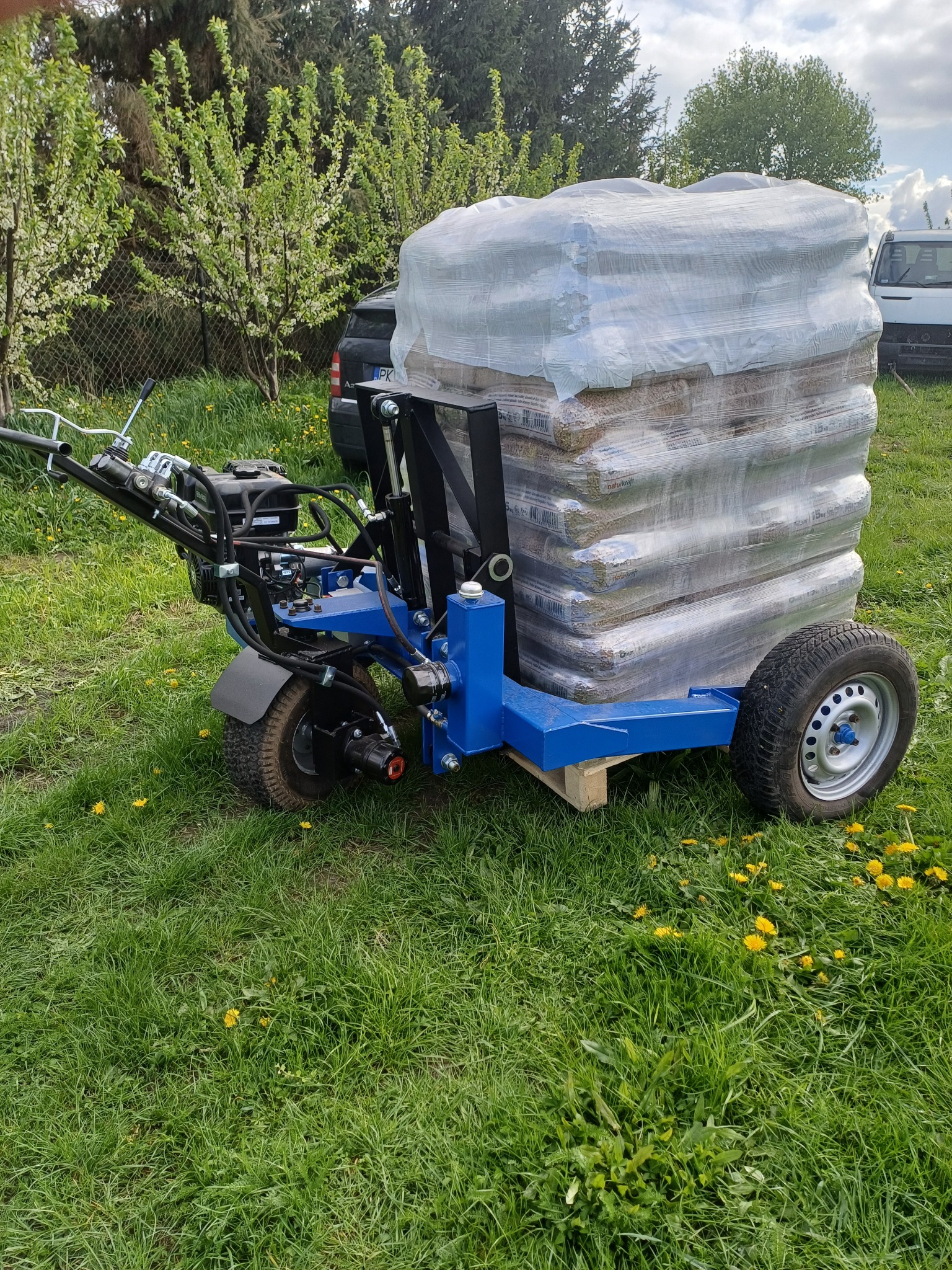 Wózek transportowy w kolorze niebieskim z załadunkiem worków, owiniętych folią, na tle zielonej trawy i kwitnących drzew.