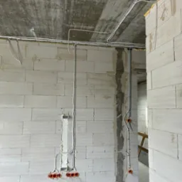 Surowy stan: białe ściany z betonu komórkowego, widoczne kable elektryczne prowadzone po ścianach i suficie. Pomarańczowe puszki elektryczne w ścianach.