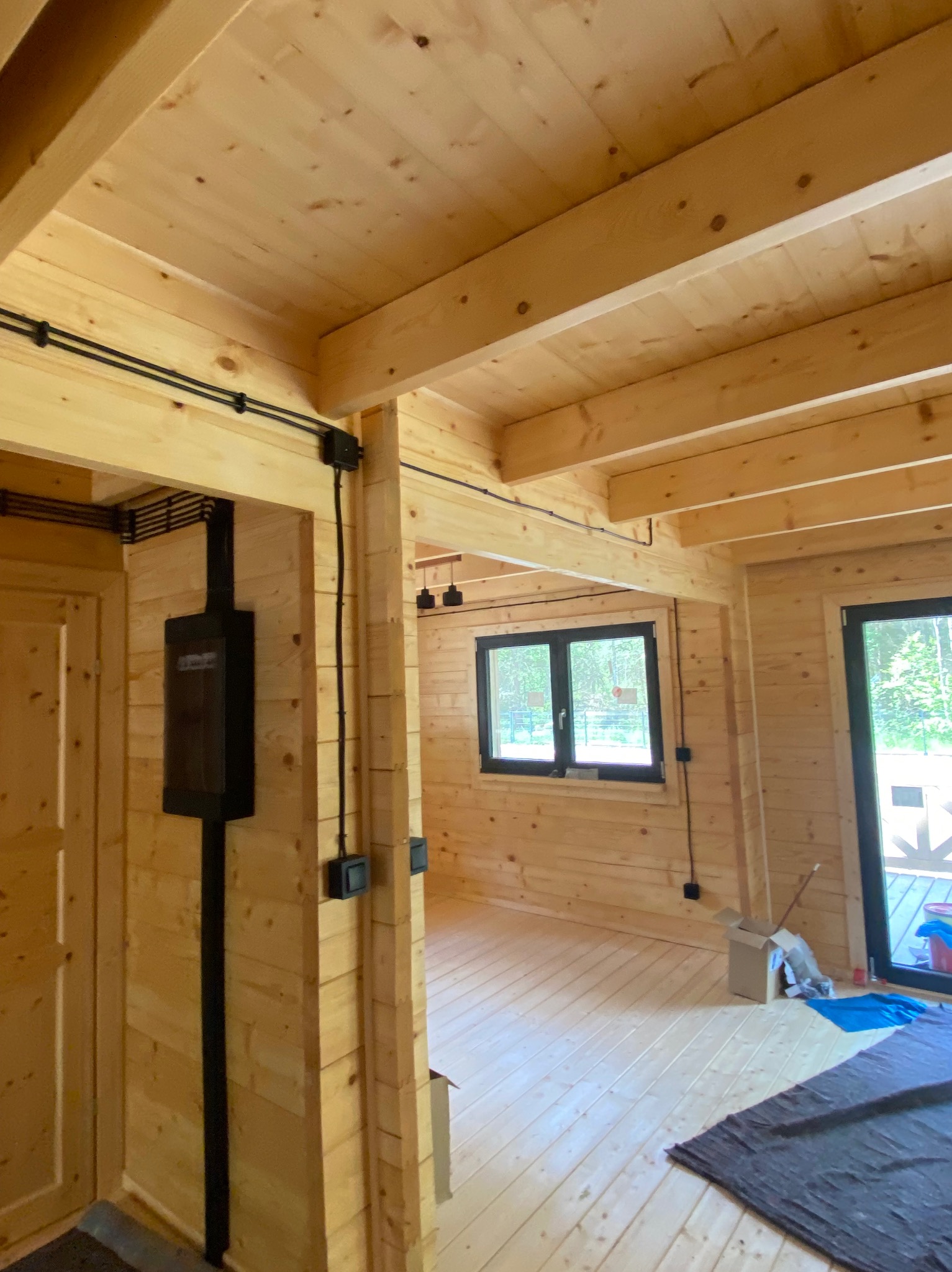 Instalacja elektryczna natynkowa w nowym, drewnianym domu. Widoczne kable w czarnych rurkach, puszki i skrzynka rozdzielcza. Świeże drewno, surowy stan.