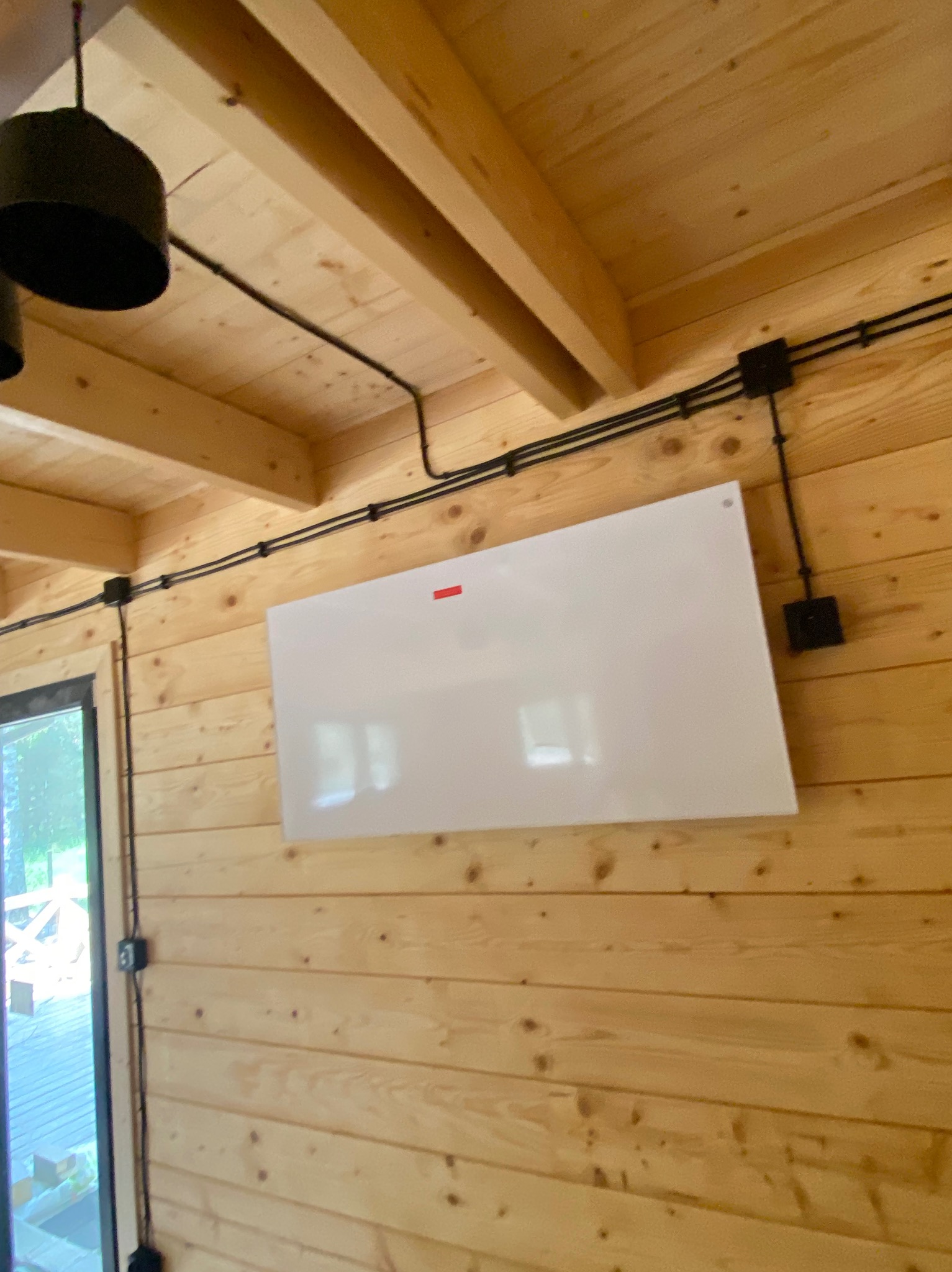 Drewniana ściana z widoczną instalacją elektryczną natynkową w czarnych peszlach i puszkach. Na ścianie zamontowany biały panel grzewczy.