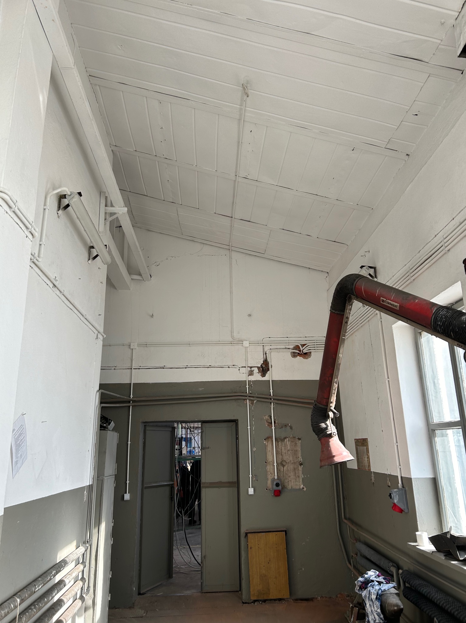 Białe rury elektryczne na ścianie i suficie w hali przemysłowej. Widoczny system wentylacyjny, okno i drzwi do kolejnego pomieszczenia.