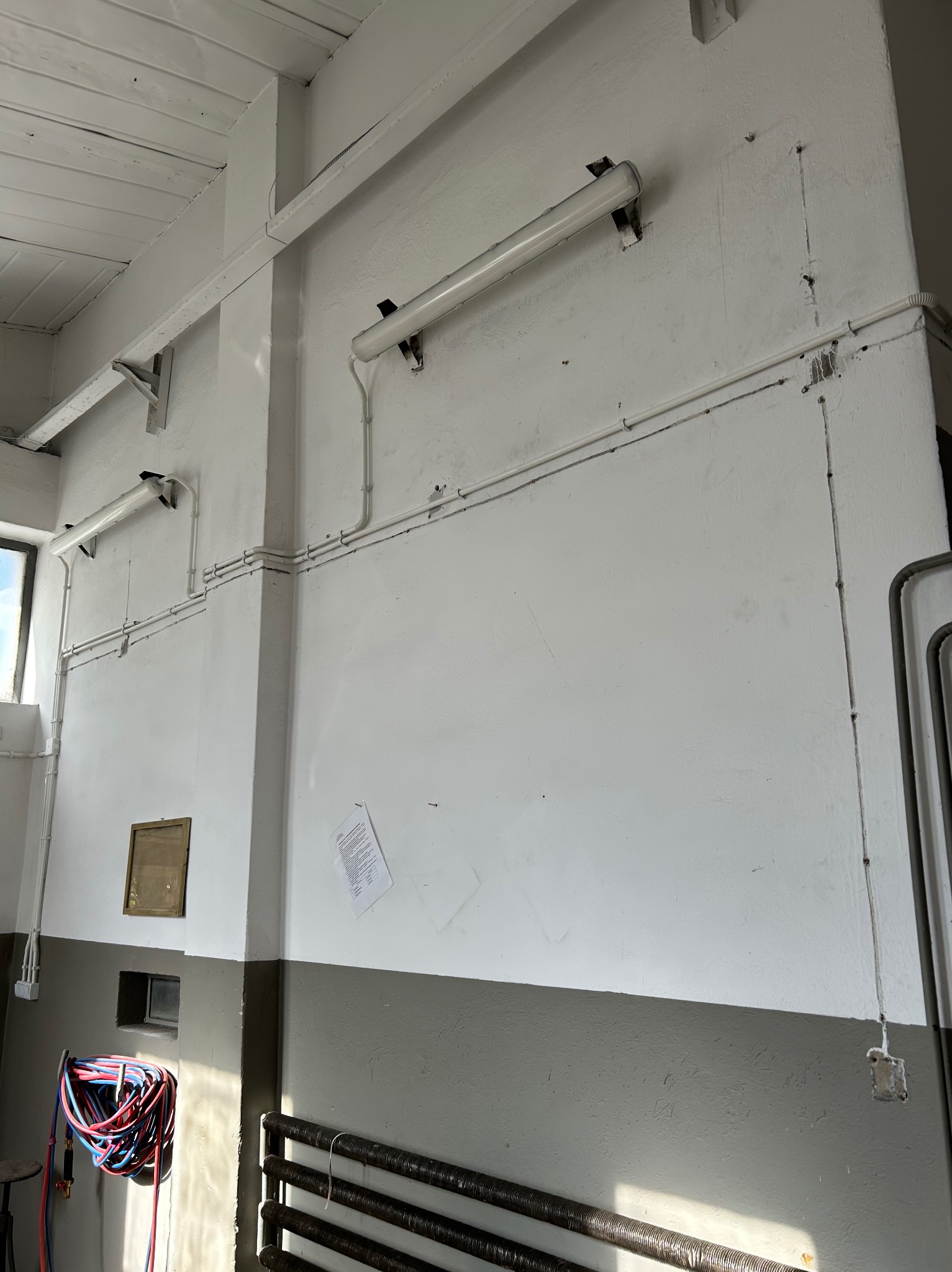 Wnętrze z widocznymi lampami fluorescencyjnymi zamontowanymi na ścianie, z widocznymi przewodami elektrycznymi. Na dole grzejnik żeberkowy.