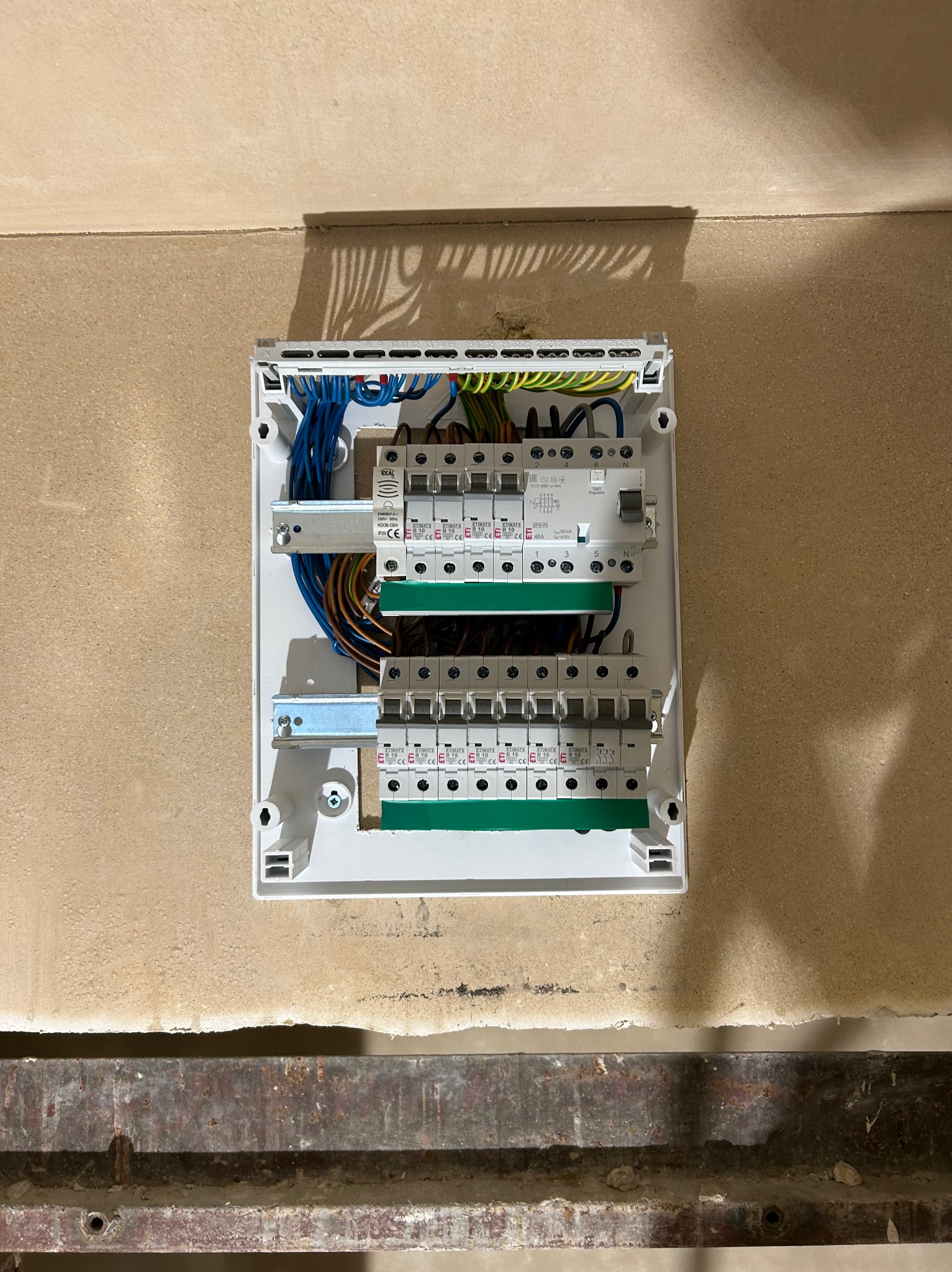 Otwarta skrzynka elektryczna z bezpiecznikami i przewodami na ścianie w surowym stanie. Widoczne elementy instalacji i zabezpieczenia, w tym wyłączniki różnicowoprądowe.