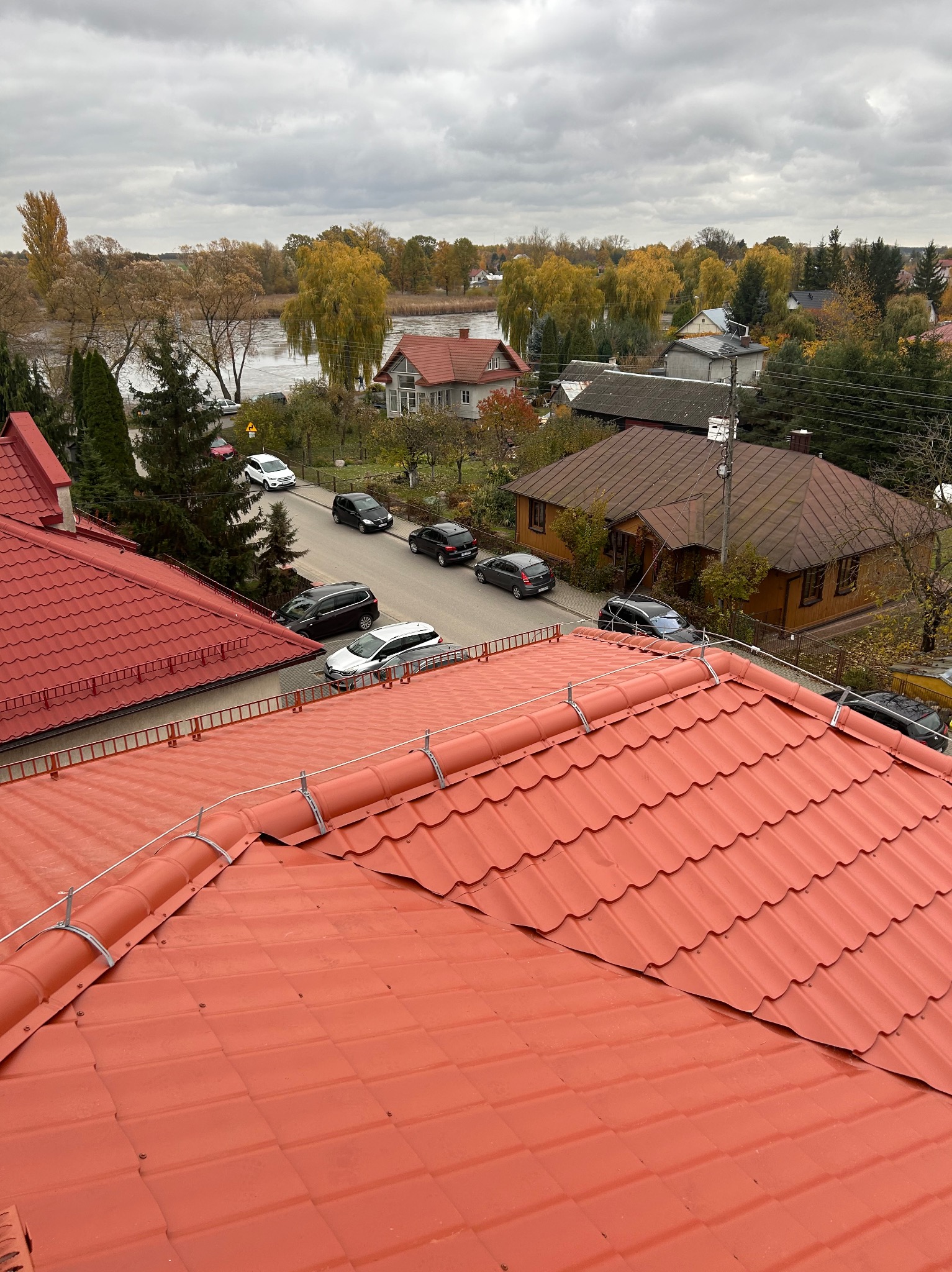 Czerwony dach z blachodachówki, zabezpieczenie odgromowe, widok z góry na okolicę z domami i drzewami w jesiennej scenerii pod pochmurnym niebem.