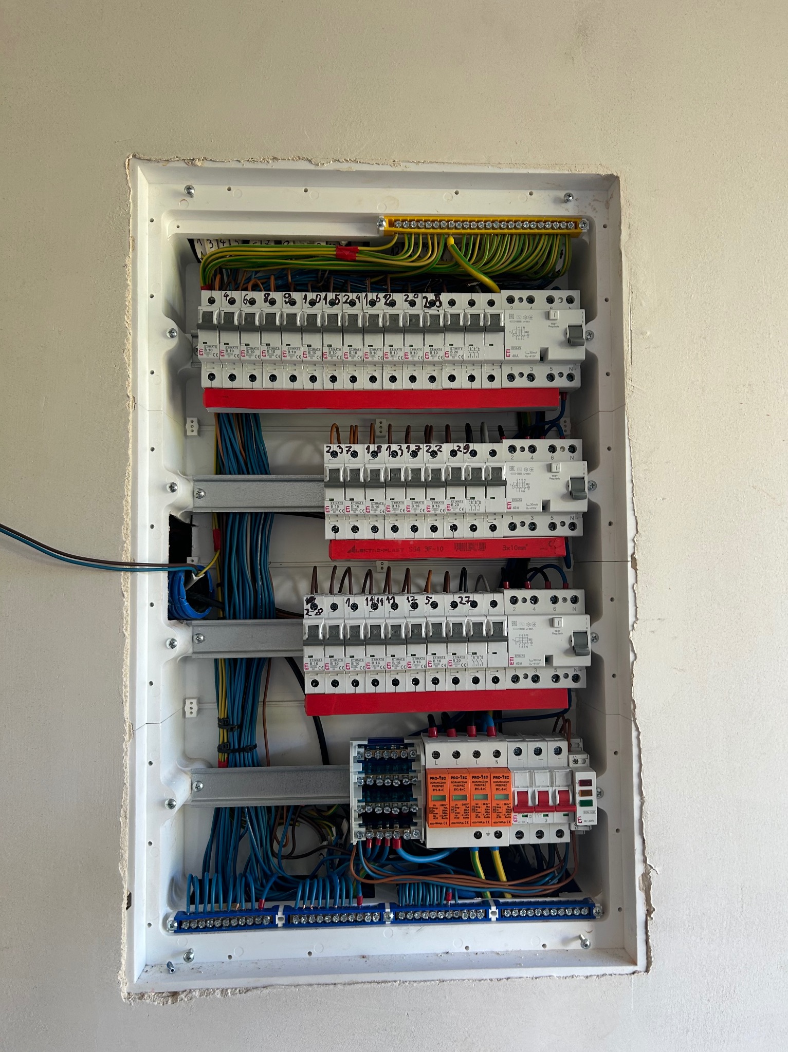 Rozdzielnica elektryczna wnękowa z zabezpieczeniami przepięciowymi i różnicowoprądowymi, widoczne przewody i listwy zaciskowe, zamontowana w surowej ścianie.