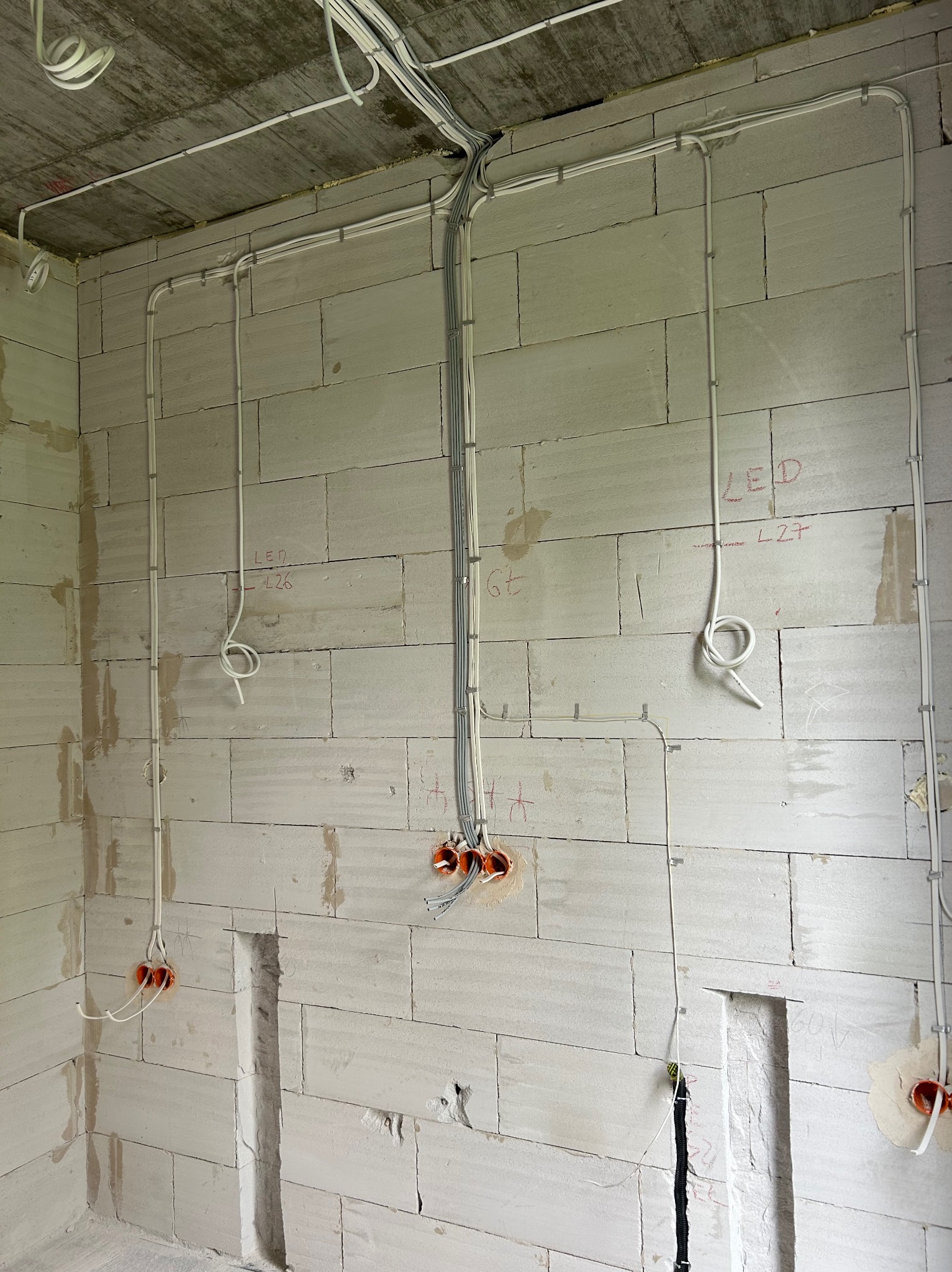 Surowa ściana z betonu komórkowego z widoczną instalacją elektryczną w peszlach i puszkach, przygotowaną do dalszych prac wykończeniowych. Widoczne oznaczenia LED.