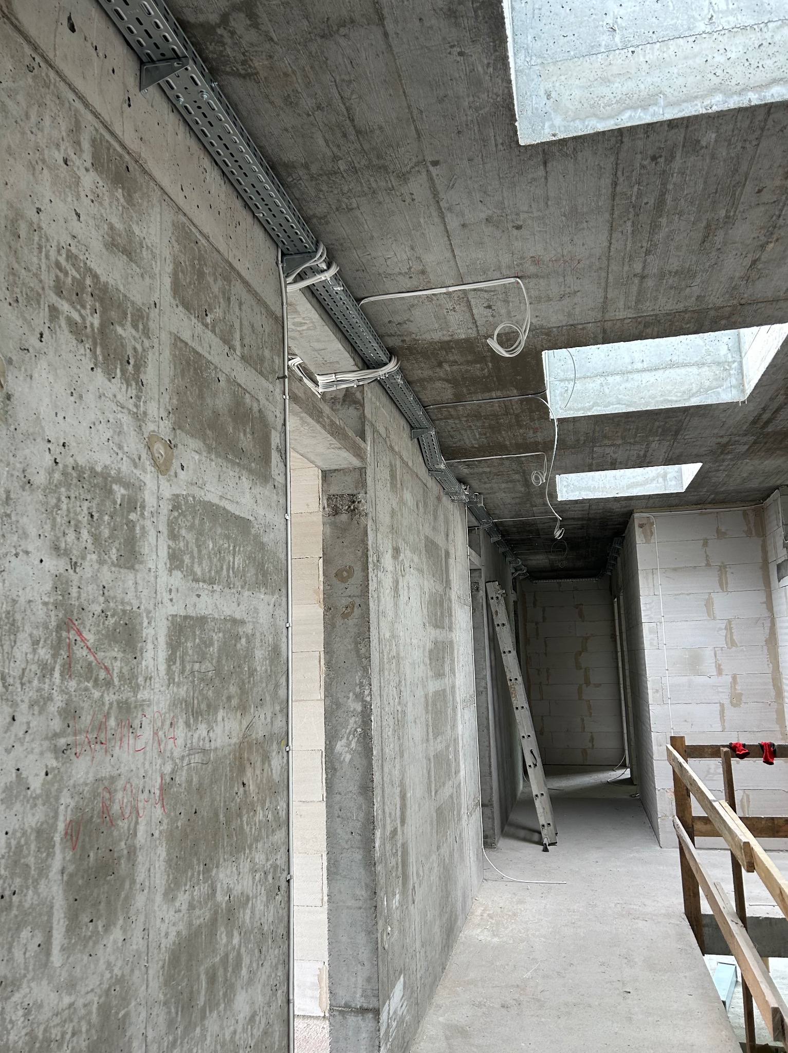 Wnętrze surowego budynku w trakcie prac. Widoczne koryta kablowe zamontowane na betonowych ścianach i suficie. W tle drabina i fragment drewnianej barierki.