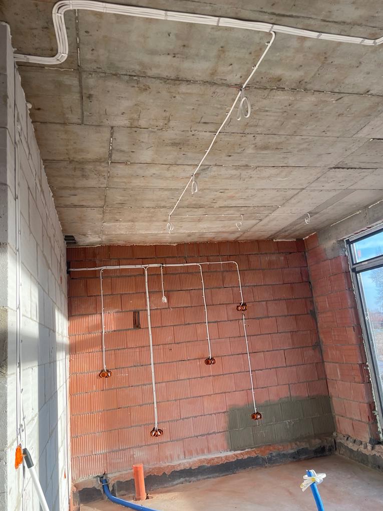 Surowy stan: instalacja elektryczna w budynku z cegły i betonu. Widoczne kable w białych listwach, puszki elektryczne w ścianie i rury na podłodze.