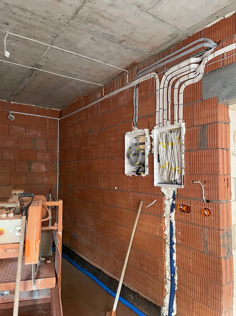 Instalacja elektryczna w surowym budynku: kable w ścianach z cegły, puszki podtynkowe, przewody ułożone w peszlach. Widoczny sprzęt budowlany.