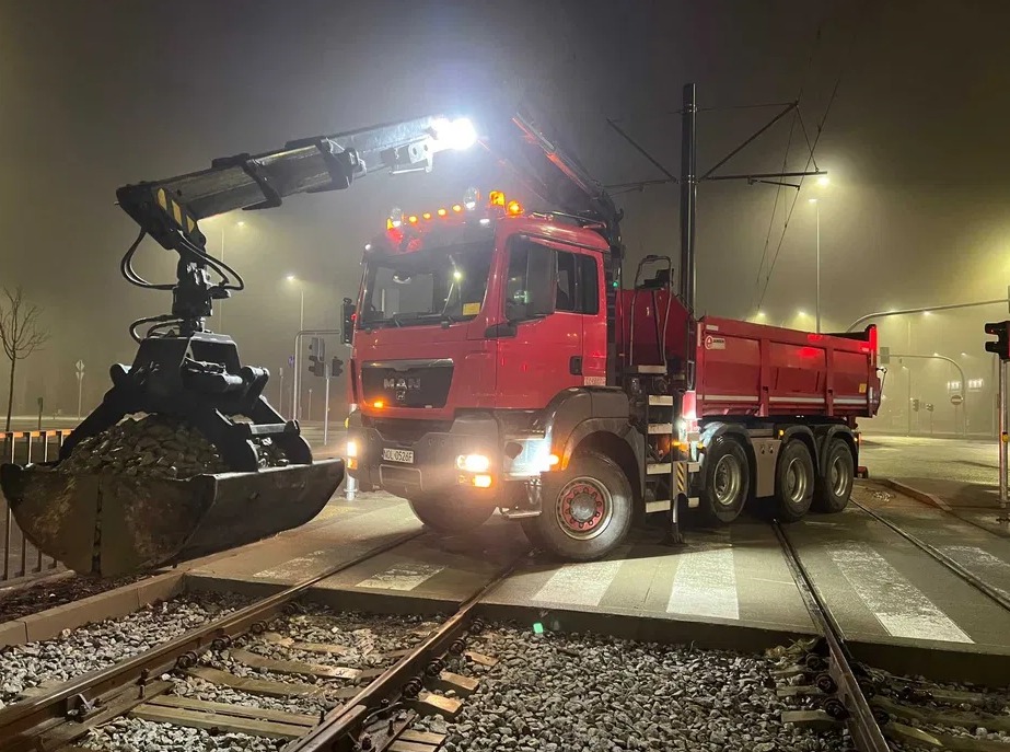 Czerwona ciężarówka MAN z chwytakiem załadowanym gruzem na torach tramwajowych w nocy, oświetlona przez reflektory. Widoczny fragment przejścia dla pieszych i sygnalizacja świetlna.