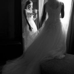 Lidiya Spirina - Czarno-białe ujęcie panny młodej w sukni ślubnej stojącej przed lustrem, w którym odbija się jej uśmiechnięta twarz i bukiet. Scena w świetle okna.