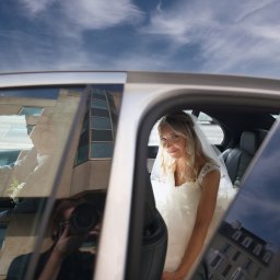 Lidiya Spirina - Uśmiechnięta panna młoda w sukni ślubnej i welonie, siedząca w luksusowym samochodzie z otwartymi drzwiami, w tle błękitne niebo i odbicie fotografa w szybie.