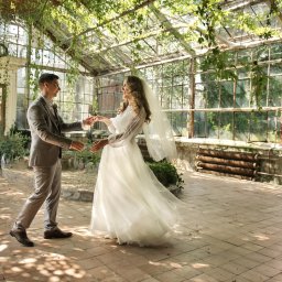 Lidiya Spirina - Para młoda w szklarni, on w szarym garniturze, ona w białej sukni z welonem. Delikatne światło, roślinność tworzą romantyczny klimat. Nietypowa lokalizacja.