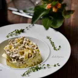 Elegancko podane tagliatelle z sosem na białym talerzu, ozdobione świeżym tymiankiem, na drewnianym stole z napojem i kwiatami w tle.