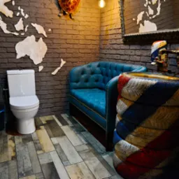 Nietypowa aranżacja toalety: ceglane ściany z mapą świata, umywalka z opon, turkusowa kanapa i drewnopodobna podłoga tworzą unikalny, industrialny klimat.