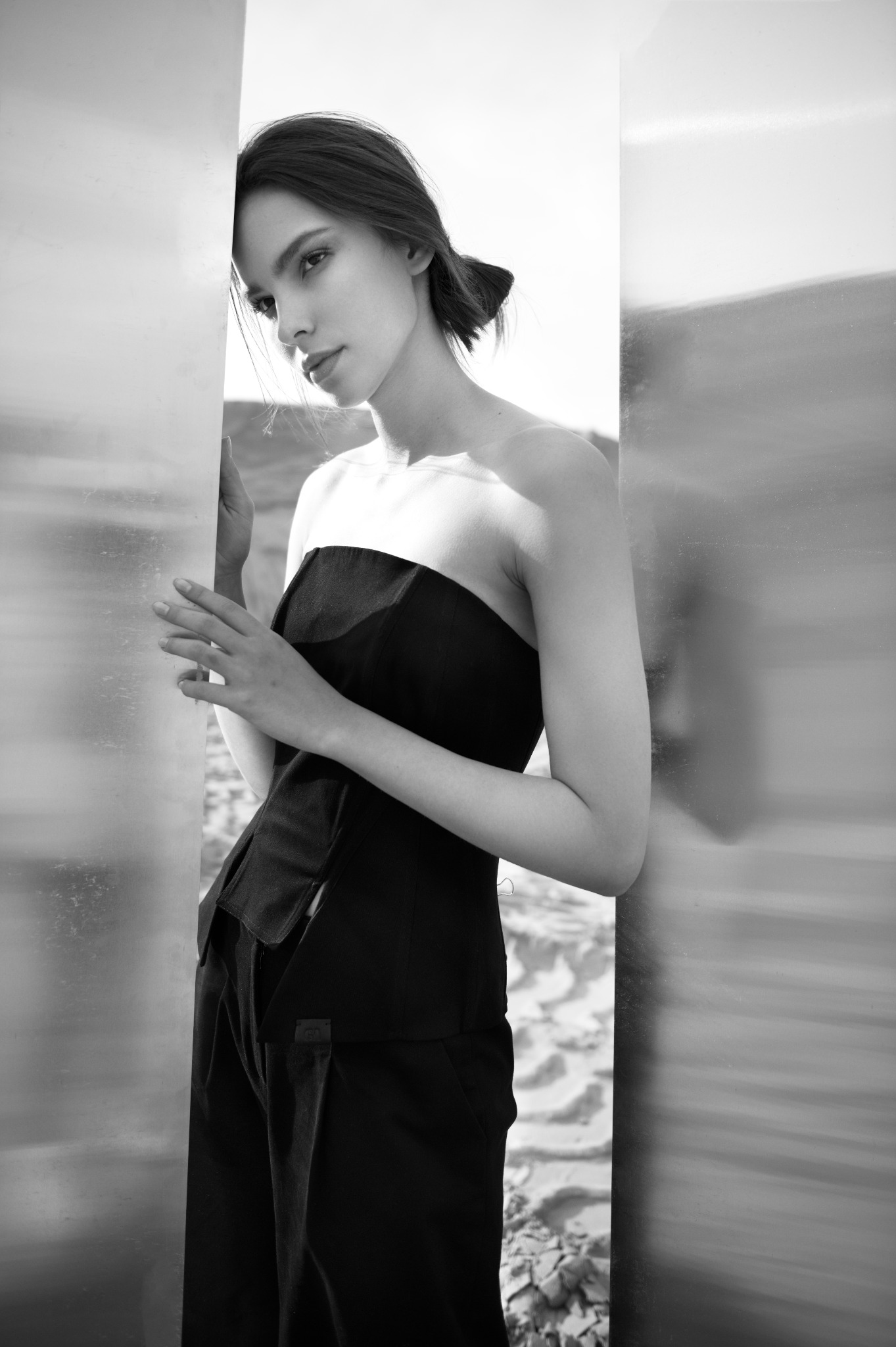 Monochromatyczne ujęcie modelki w czarnym gorsecie i spodniach, opierającej się o metaliczną powierzchnię, z delikatnym makijażem i upiętymi włosami, w naturalnym świetle.