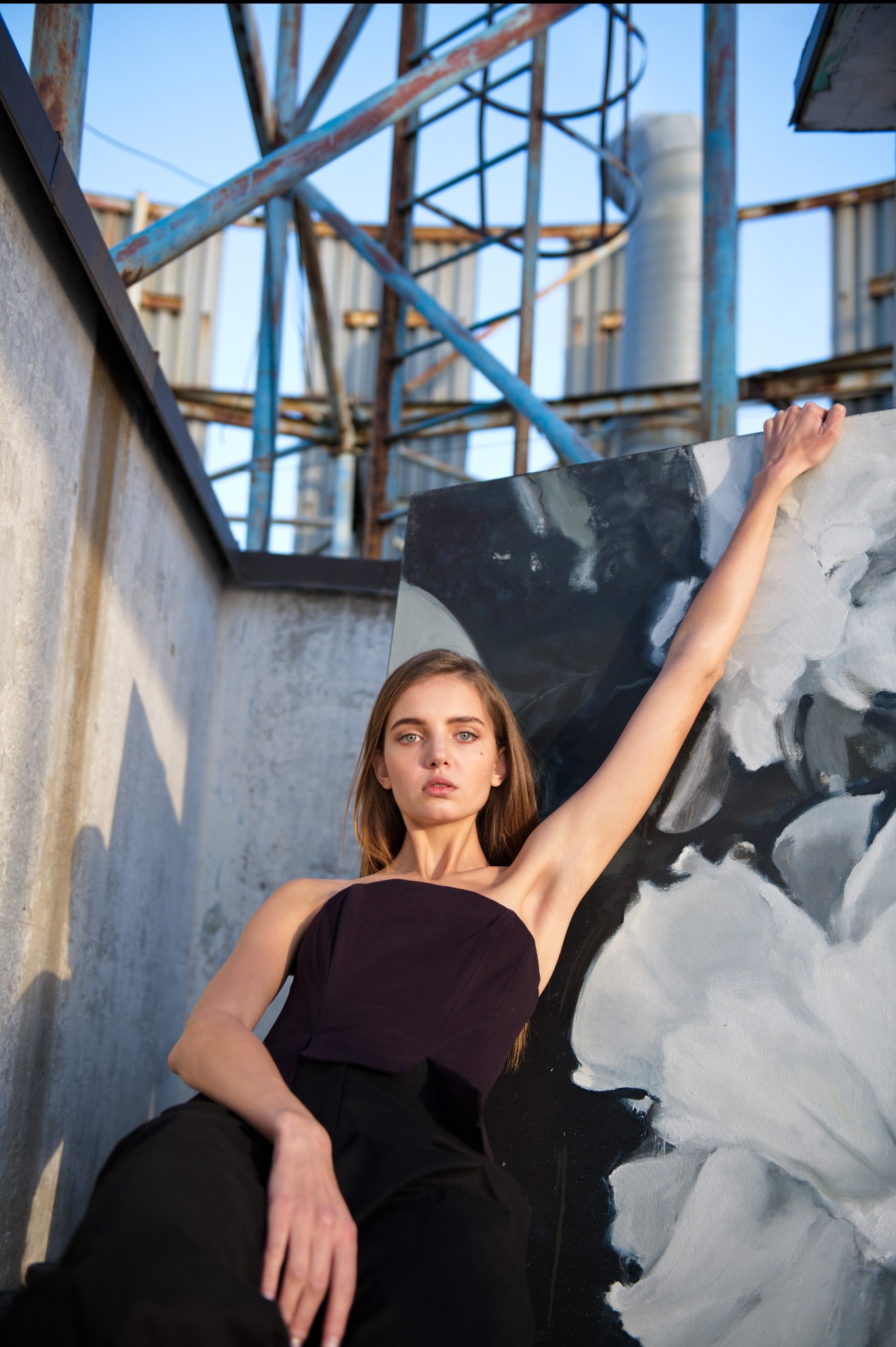 Portret kobiety z obrazem na tle metalowej konstrukcji i błękitnego nieba. Modelka w czarnym stroju, wyciągnięta ręka, artystyczna poza, naturalne światło.