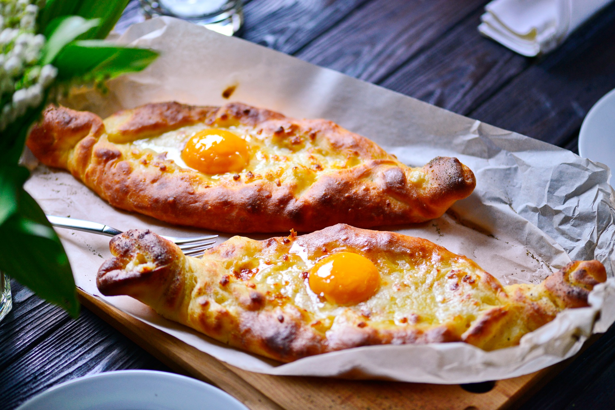 Dwa chaczapuri adżarskie z jajkiem na papierze do pieczenia, leżące na drewnianej desce. W tle ciemne, drewniane deski, kwiaty w wazonie i serwetka.