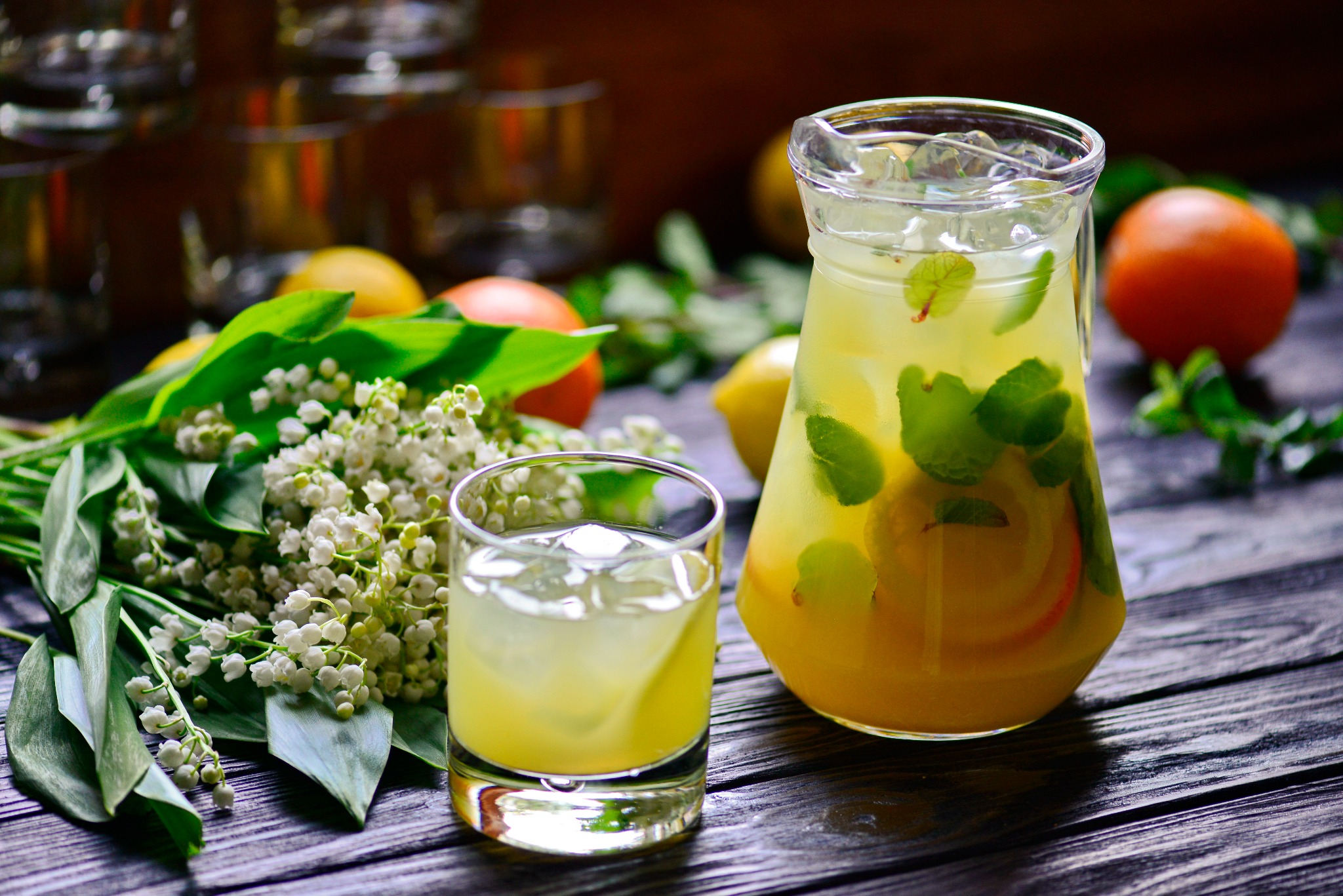 Szklanka i dzbanek lemoniady z lodem, miętą i owocami na ciemnym drewnianym stole, obok konwalie i cytrusy. Naturalne światło, ciepła kolorystyka, rustykalny charakter.