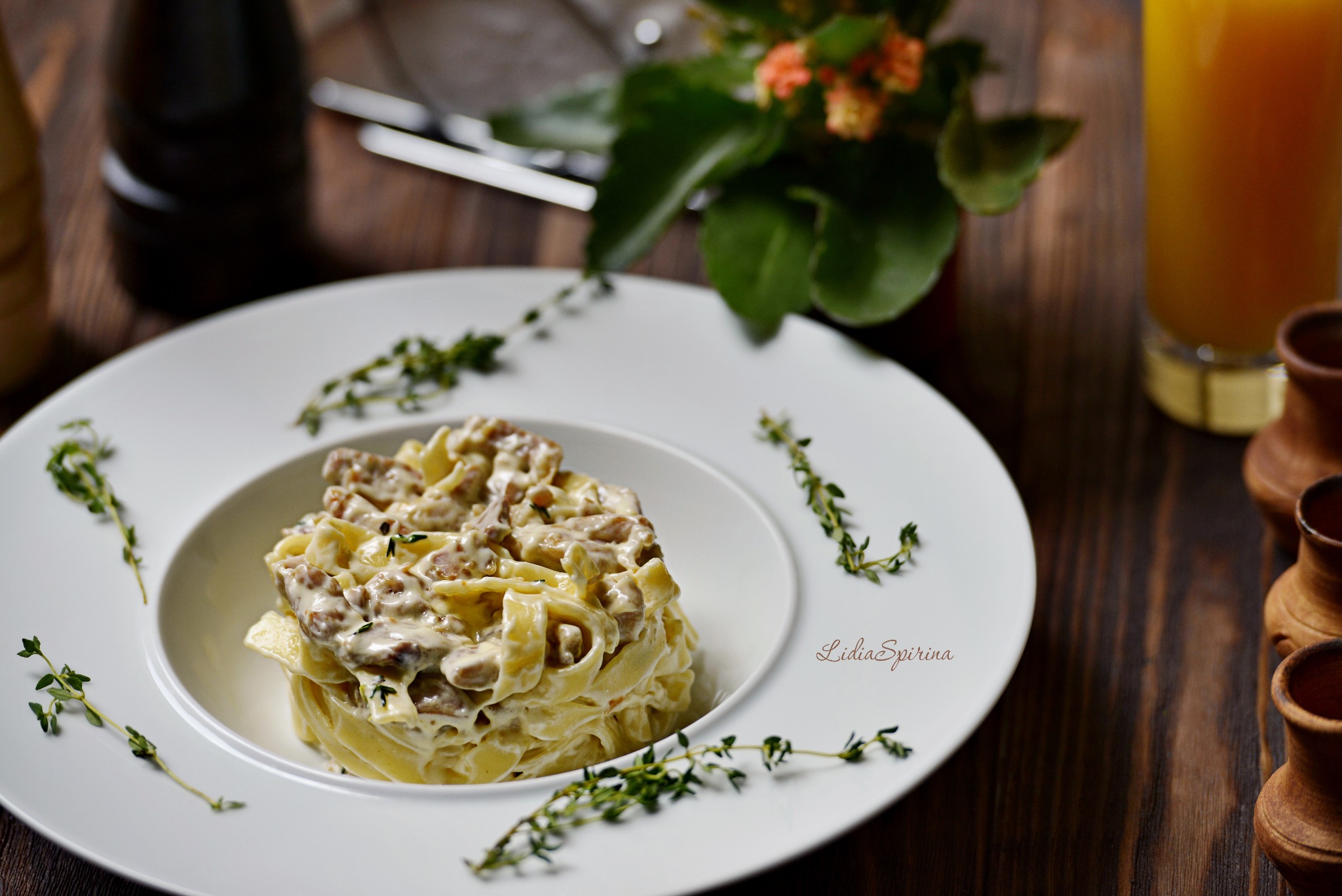 Elegancko podane tagliatelle z sosem na białym talerzu, ozdobione świeżym tymiankiem, na drewnianym stole z napojem i kwiatami w tle.
