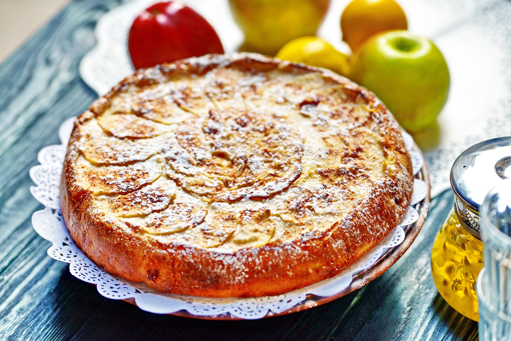 Apetyczna szarlotka posypana cukrem pudrem, na koronkowej serwetce, z jabłkami i imbrykiem herbaty w tle, tworząca ciepłą, domową atmosferę.