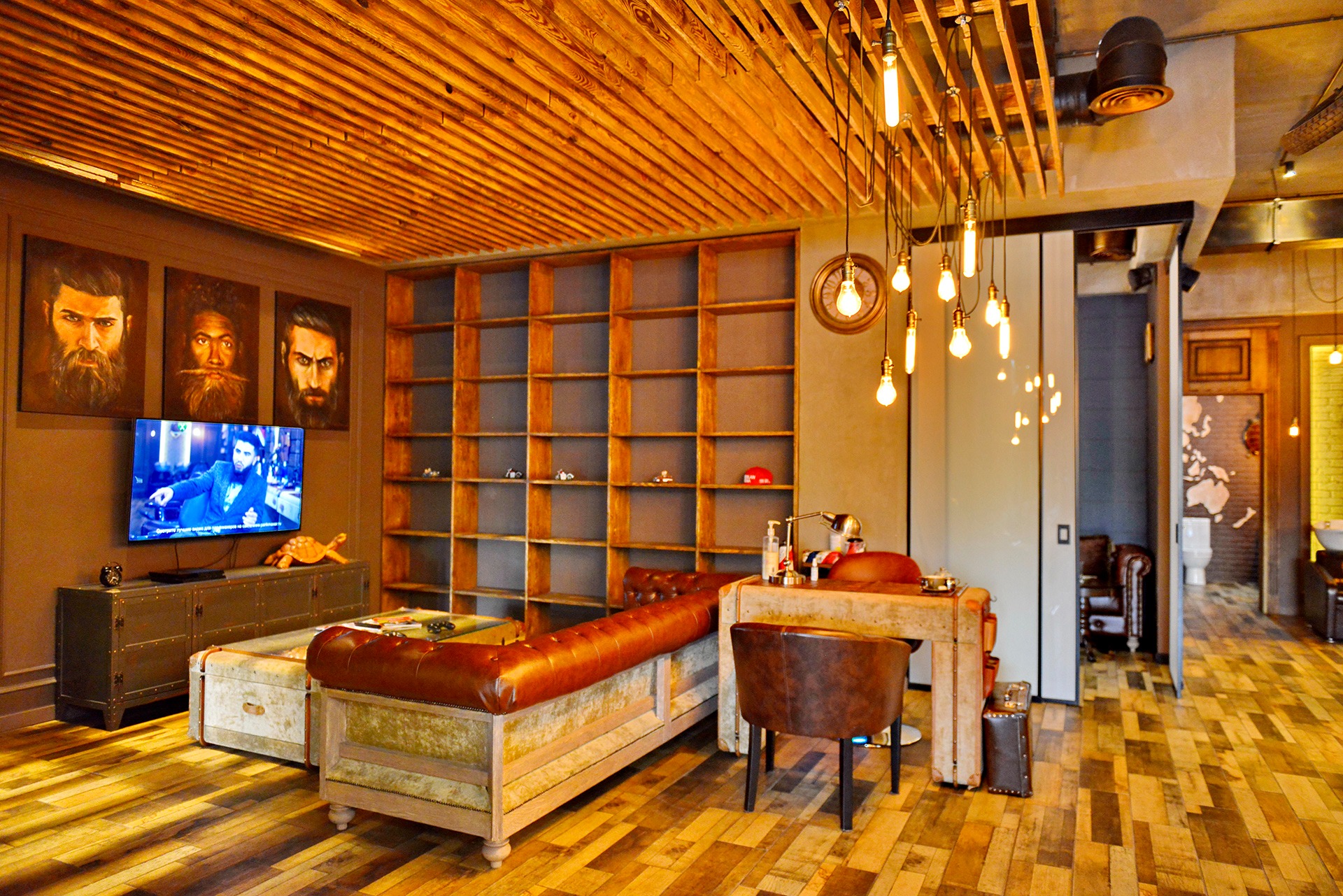 Wnętrze salonu barberskiego w stylu loft: drewniany sufit, skórzana kanapa, regał z modelami aut, obrazy brodatych mężczyzn i dekoracyjne oświetlenie.
