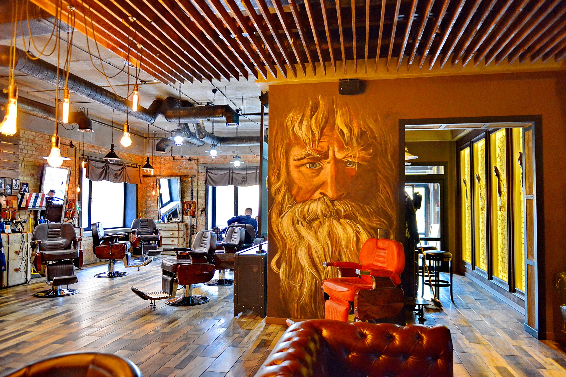 Wnętrze barber shopu z muralem brodatego mężczyzny na ścianie, skórzanymi fotelami fryzjerskimi i drewnianą podłogą. Widoczne industrialne oświetlenie i ceglane ściany.