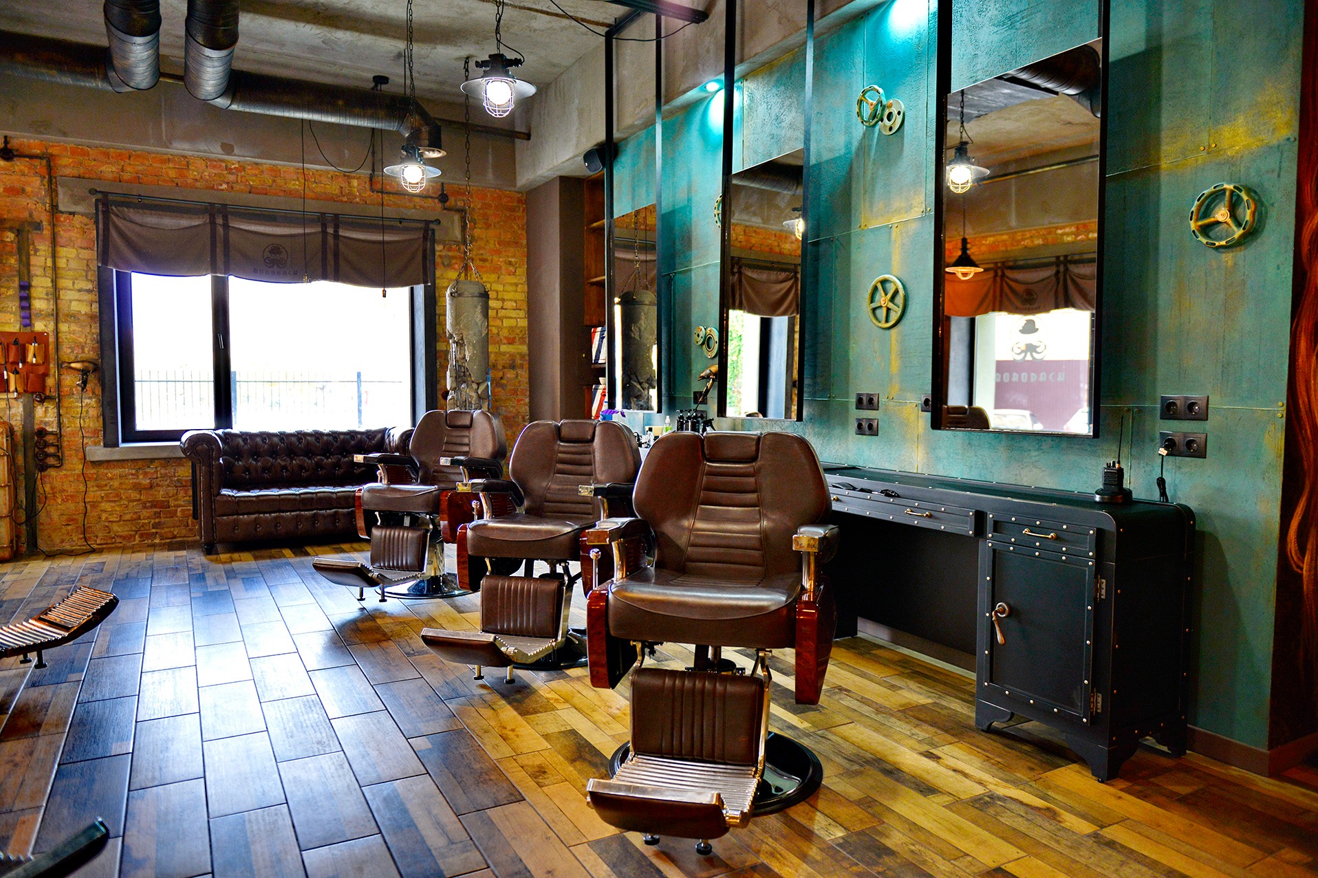 Wnętrze barber shopu w stylu industrialnym: trzy skórzane fotele fryzjerskie, ceglana ściana, metalowe lampy i lustra z dekoracjami w stylu steampunk.