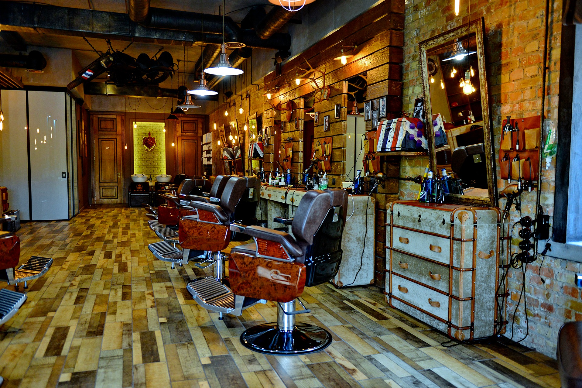 Wnętrze barber shopu w stylu retro: drewniana podłoga, ceglane ściany, skórzane fotele fryzjerskie i oświetlenie żarówkowe. Widoczne detale i akcesoria fryzjerskie.