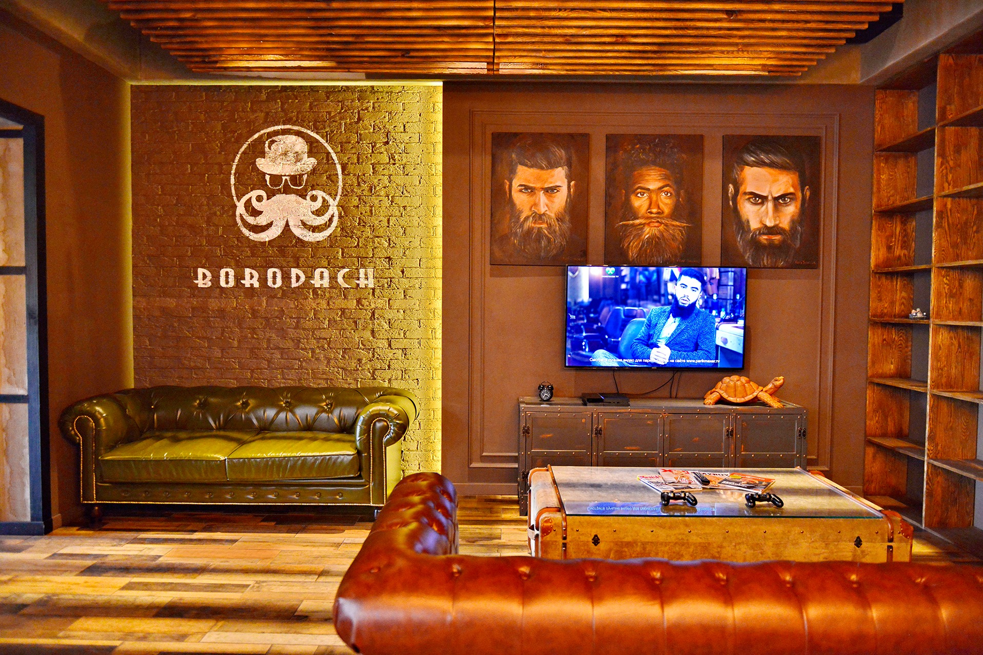Wnętrze salonu barberskiego z ceglaną ścianą, logo z wąsami, skórzanymi kanapami, obrazami brodatych mężczyzn i telewizorem. Ciepłe oświetlenie i drewniane elementy.