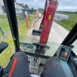 Podłącz PROjekt Bartłomiej Zaręba - Widok z kabiny koparki na wykop wzdłuż ogrodzenia, z widocznym czerwoną rurą i stertą kostki brukowej w tle, sugerujący prace ziemne pod instalacje.