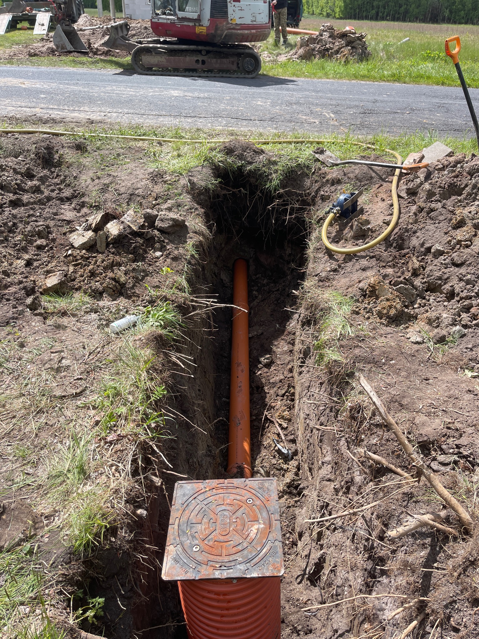 Pomarańczowa rura kanalizacyjna w wykopie ziemnym z widoczną studzienką kanalizacyjną i sprzętem budowlanym w tle, sugerująca etap prac instalacyjnych.