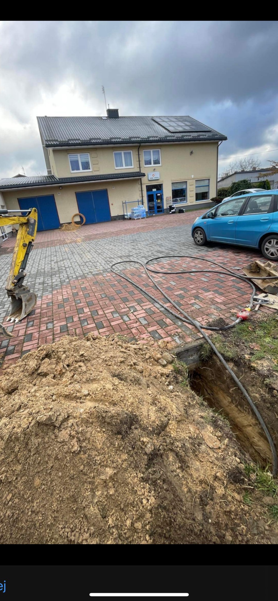 Wykop z ułożonymi rurami na posesji z budynkiem i niebieskim autem w tle. Żółta mini koparka obok. Prace ziemne związane z instalacją.