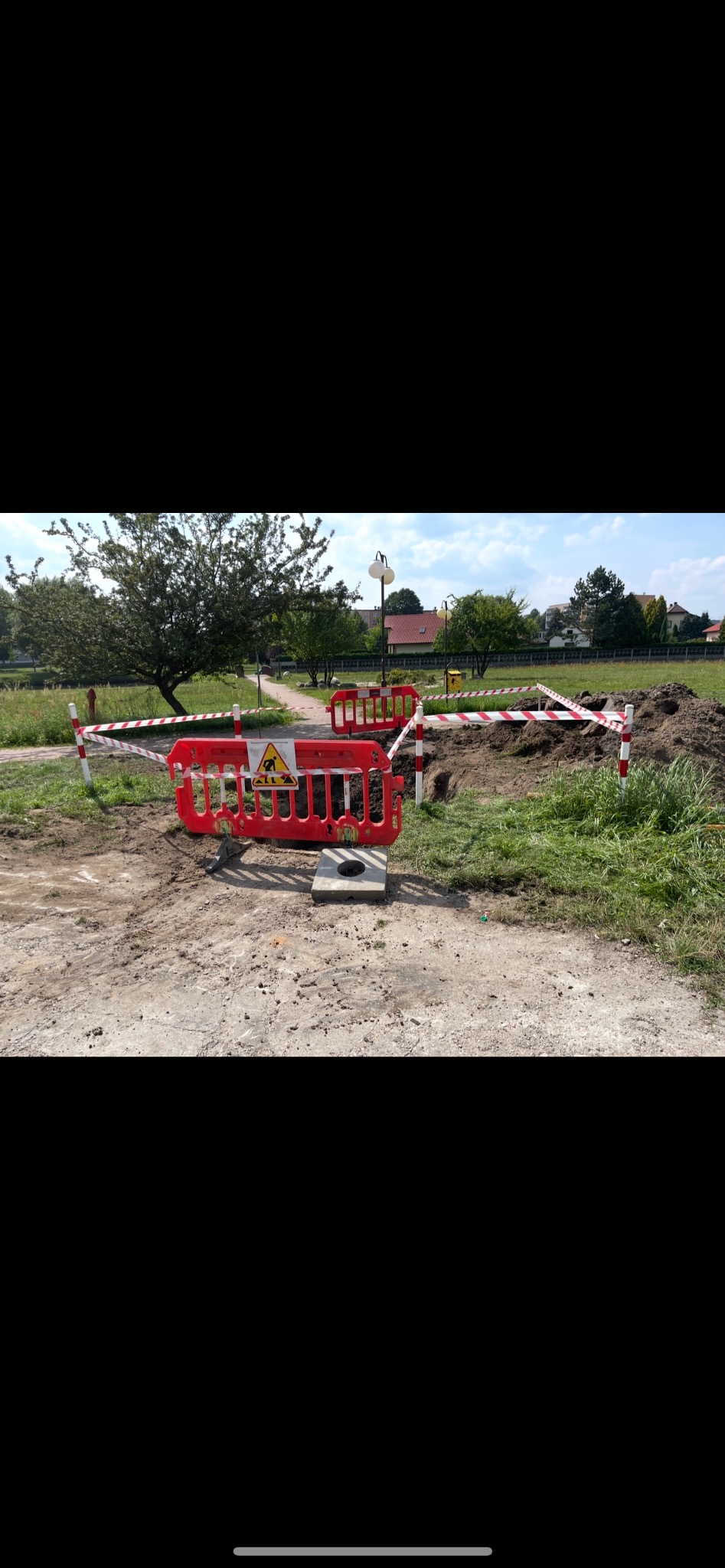 Wykonany wykop zabezpieczony czerwoną barierką i taśmą ostrzegawczą na terenie zielonym z widocznymi budynkami w tle. Prace ziemne zabezpieczone.