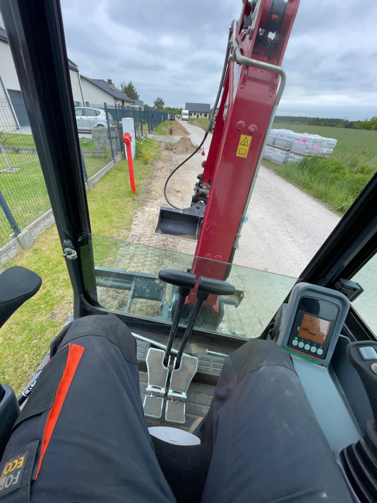 Widok z kabiny koparki na wykop wzdłuż ogrodzenia, z widocznym czerwoną rurą i stertą kostki brukowej w tle, sugerujący prace ziemne pod instalacje.