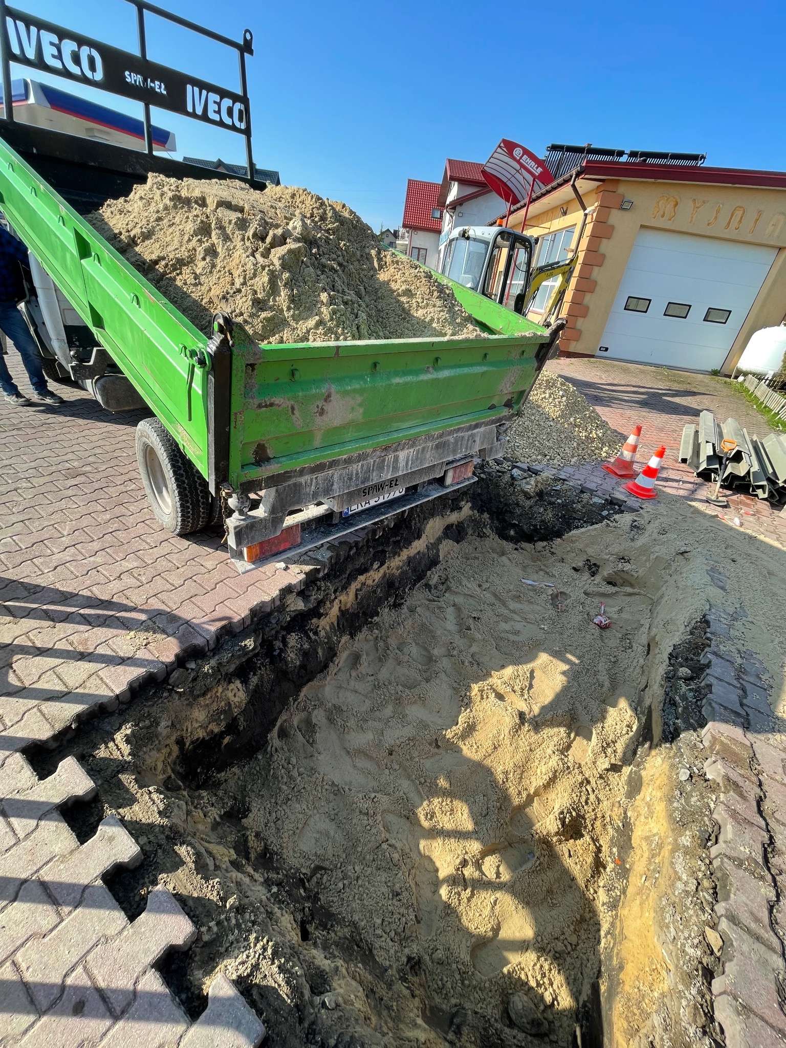 Zielona wywrotka Iveco z piaskiem na tle wykopu w kostce brukowej, pomarańczowe pachołki i budynek w tle. Realizacja przyłącza wod-kan na posesji.