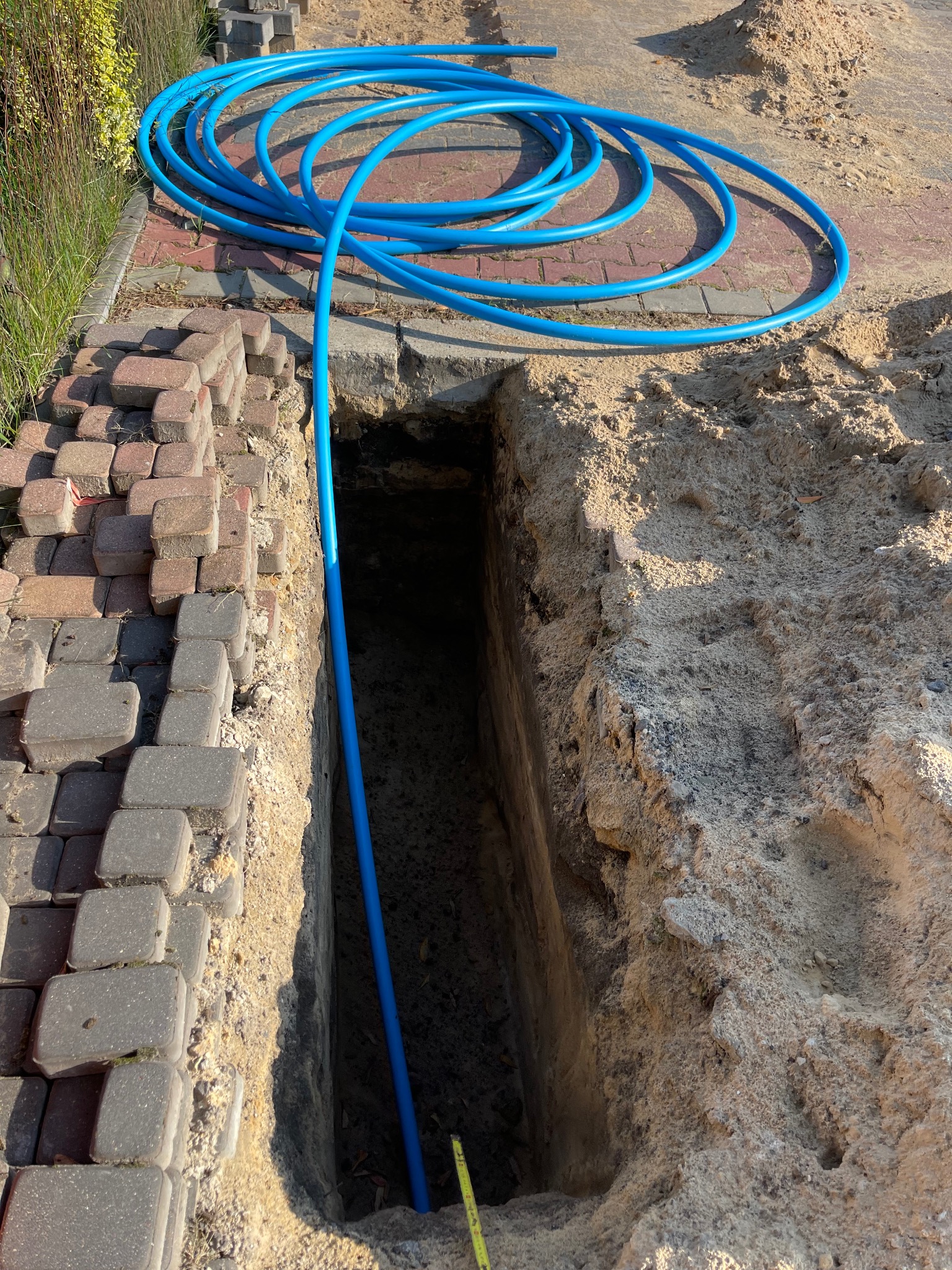 Niebieska rura wodociągowa w wykopie ziemnym, zwoje rury leżą na kostce brukowej. Widoczny fragment trawnika i kostki brukowej. Instalacja przyłącza wodociągowego.