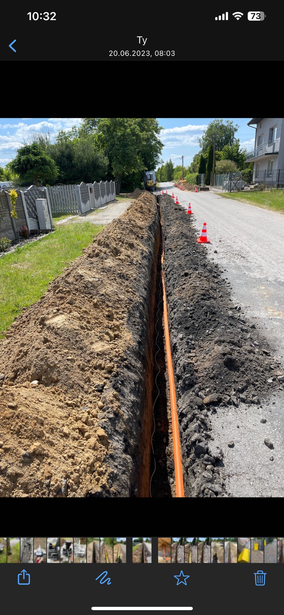 Wykop z pomarańczową rurą w ziemi, zabezpieczony stożkami na drodze. Widoczna koparka w oddali. Prace ziemne w słoneczny dzień.