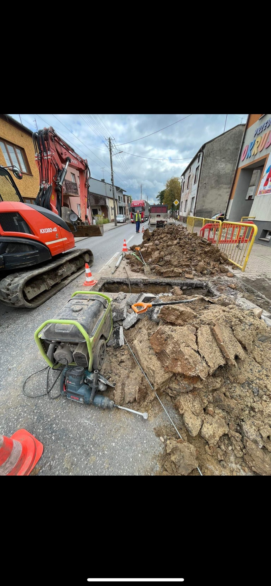 Wykonanie wykopu pod przyłącze wod-kan na ulicy, z użyciem koparki Kubota KX080-4a i agregatu prądotwórczego Bosch. Widoczne zabezpieczenia i robotnicy.