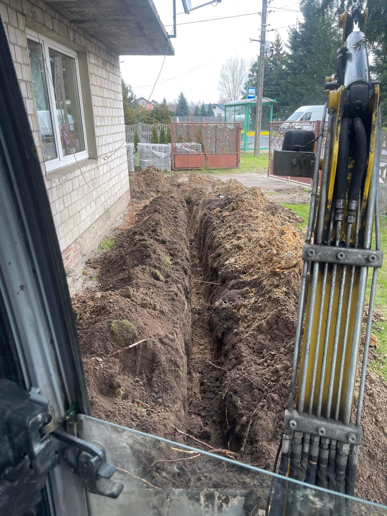 Wykopany rów w ziemi, widok z kabiny koparki, w tle budynek z białej cegły i przystanek autobusowy. Przygotowanie do instalacji przyłącza wod-kan.
